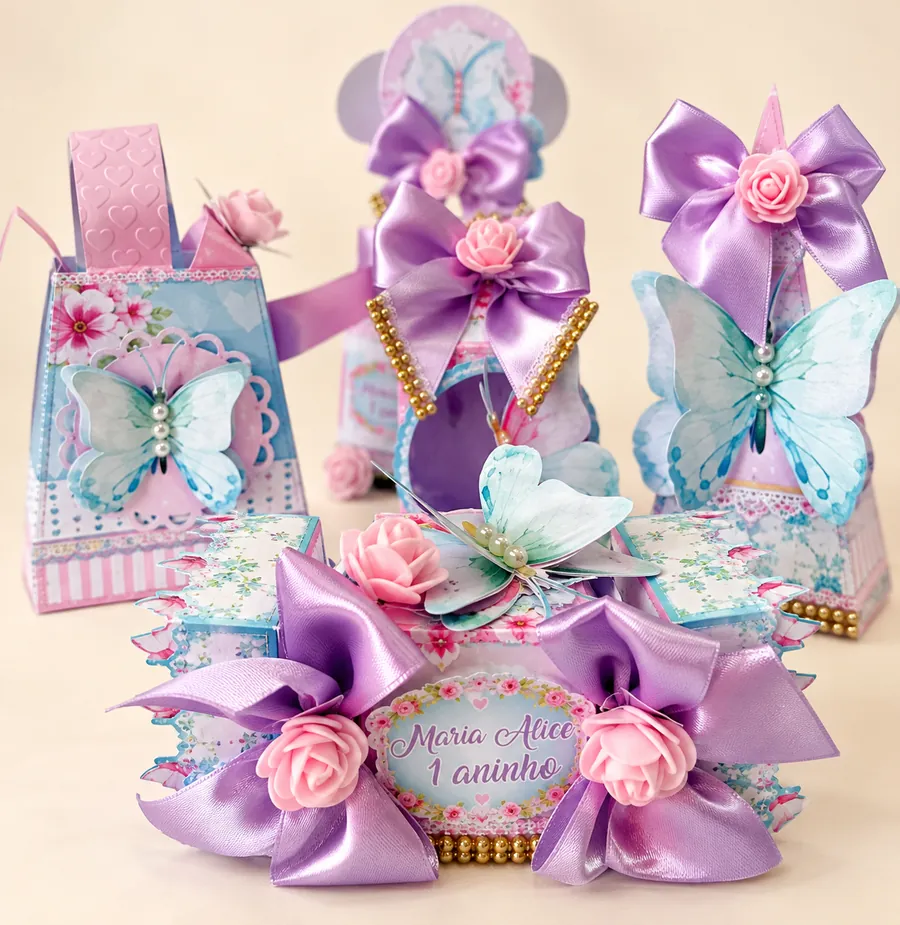 Conjunto de lembrancinhas em papel com estampa floral azul e rosa, borboletas, laços de cetim lilás e rosas, com a etiqueta Maria Alice 1 aninho.